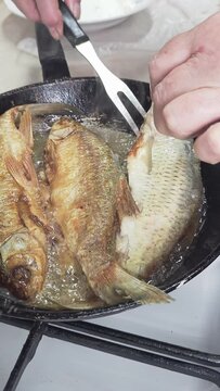 woman hands flipping with a carving fork whole vobla fish while frying in a cast iron skillet with oil at domestic kitchen.