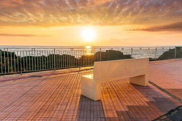 beautiful white stone bench on seafront embarkment during beautiful sunrise or sunset with sidewalk with pavement, sea surf and nice cloudy sunset sky