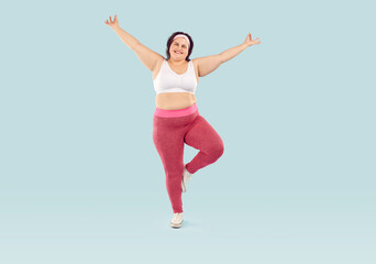 Fototapeta na wymiar Happy cheerful joyful fat overweight woman in white sports bra and pink yoga pants isolated on light blue background enjoying fitness workout, doing exercise, standing on one leg. Weight loss concept
