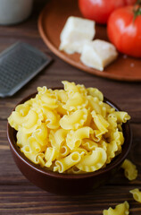 Raw Creste Di Gallo pasta in a ceramic bowl with fresh tomatoes and parmesan cheese on a brown wooden board. Rooster comb. The concept of traditional Italian cuisine. Vertical orientation.