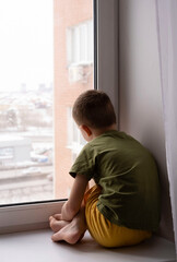 Sad cute child looks out window and feels sad. Boy is depressed. Vertical image