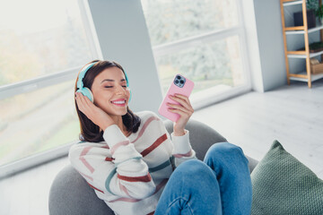 Photo of adorable cheerful girl sit chair closed eyes hold smart phone touch headphones listen...