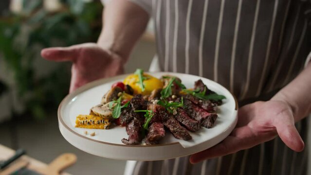 Cropped shot of unrecognizable male chef demonstrating white plate with sliced beef steak and grilled vegetables decorated with pine nuts and microgreen
