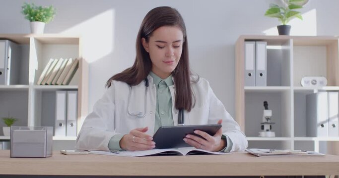 Satisfied female doctor smiling dreamily while working with digital tablet in hospital office. Young Caucasian doctor in white coat scrolls something on tablet and smiles dreamily looking away.