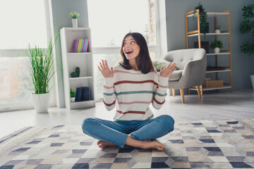 Full body photo of impressed positive lady sit barefoot carpet floor raise hands look away unbelievable reaction flat indoors