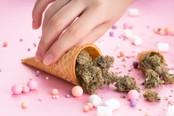 Dry buds of medical marijuana lie in ice cream waffle cones, a woman's hand holds on a pink background. There are candies and marshmallows around. Alternative medical treatment with cannabis
