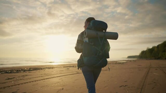 Woman tourist walks sea beach at sunset. Female traveler carrying big backpack with touristic equipment, back view. Hiker enjoying admiring nature. Travel tourism relaxed, vacations activity concept.
