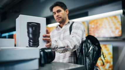 Fototapeta na wymiar Handsome Young Man Shopping for a Camera Lens in a Home Electronics Store. Multiethnic Customer Comparing Different Photo Lens Features, Sale Prices and Image Quality in a Photography Shop