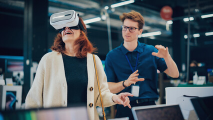Senior Customer Talking with Retail Electronics Shop Salesperson while Choosing a VR Headset as a...