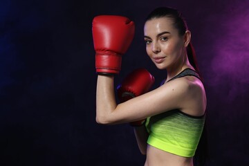 Portrait of beautiful woman wearing boxing gloves training in color lights and smoke on black background. Space for text