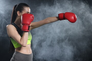 Woman wearing boxing gloves training in smoke on black background. Space for text