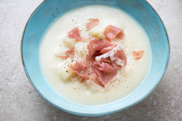 Middle closeup of potato soup served with prosciutto in a turquoise bowl, horizontal shot on a beige stone surface, selective focus