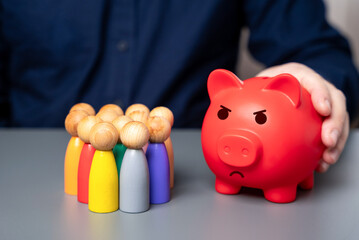 A businessman holds a red piggy bank next to a group of people figures. Lack of budget funds. Poor...