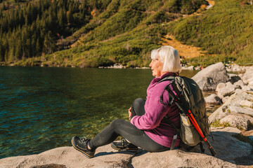 Rear shot of traveler 60s woman influencer in sportswear with backpack sitting on the rocky shore...