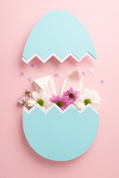 Appreciate our deluxe Easter display. Top view vertical photo of delicate chrysanthemums, confetti and bunny ears emerging from a blue eggshell on a pastel pink setting