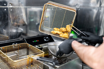 A person in black gloves is frying chicken pieces in a commercial kitchen using a metal strainer. Steam rises as the chicken cooks