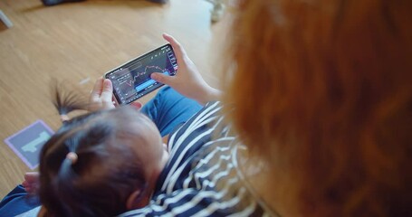 A woman trades stocks on a mobile device while caring for an infant at home - Powered by Adobe
