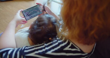 A woman trades stocks on a mobile device while caring for an infant at home - Powered by Adobe