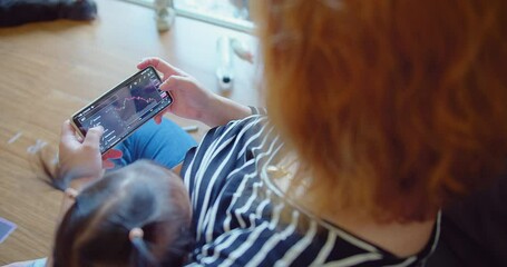 A woman trades stocks on a mobile device while caring for an infant at home - Powered by Adobe