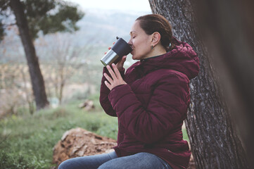 Happy relaxed young woman drinking hot tea from a thermos mug in the early spring forest