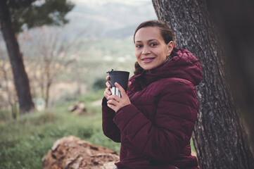 Young woman hiker traveler drinks tea in the nature, sitting on log in early spring forest, smiling looking at camera