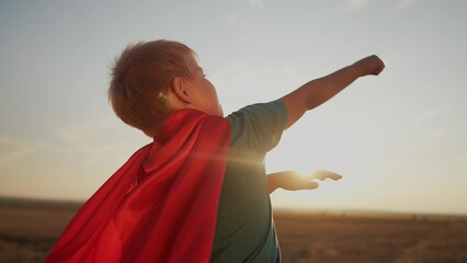 Fototapeta na wymiar boy with the cloak. concept of a happy childhood and family for a child. a child in a superhero costume stretched his hand forward, a red cape and a blue T-shirt, sunset on the background lifestyle
