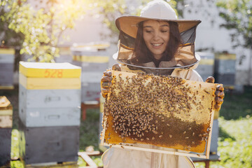 Happy smiling female Beekeeper in protective suit holding honeybee frame with bees at apiary