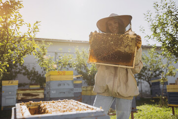 Happy smiling female Beekeeper in protective suit holding honeybee frame with bees at apiary