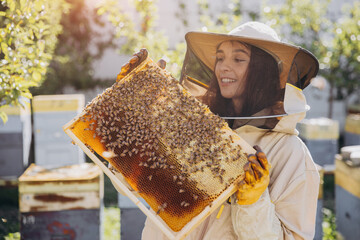 Happy smiling female Beekeeper in protective suit holding honeybee frame with bees at apiary