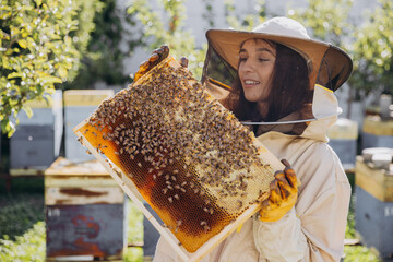 Happy smiling female Beekeeper in protective suit holding honeybee frame with bees at apiary