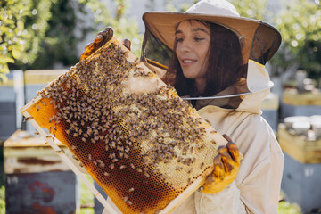 Happy smiling female Beekeeper in protective suit holding honeybee frame with bees at apiary