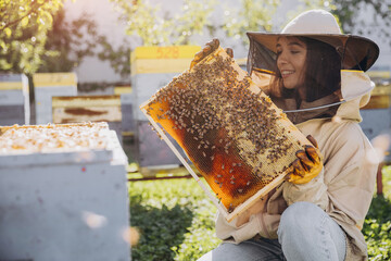 Happy smiling female Beekeeper in protective suit holding honeybee frame with bees at apiary