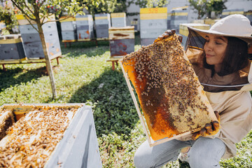 Happy smiling female Beekeeper in protective suit holding honeybee frame with bees at apiary