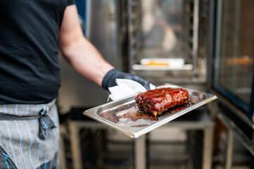 A chef in a professional kitchen holds a tray of freshly cooked, juicy ribs. The ribs are glazed and tender, with stainless steel kitchen equipment in the background