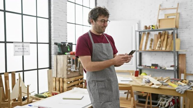 A young hispanic man in a workshop smiles while using a smartphone, surrounded by carpentry tools and woodworks.