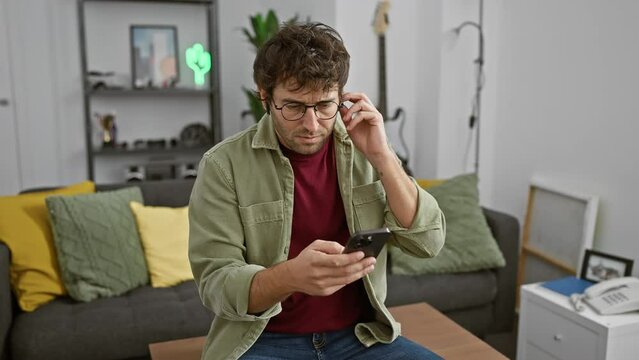 A young hispanic man with headphones and glasses using a smartphone at home, exuding a casual and focused atmosphere.
