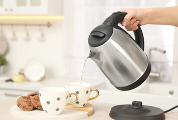 Woman pouring hot water from electric kettle into cup in kitchen, closeup. Space for text