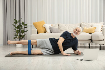 Senior man doing exercise with fitness elastic band near laptop on mat at home
