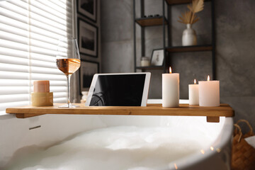 Wooden tray with tablet, wine, candles and toiletries on bathtub in bathroom