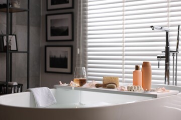 Wooden tray with wine, toiletries and flower petals on bathtub in bathroom