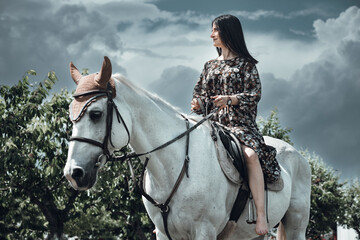 Woman in floral dress riding a white horse beneath the dramatic cloud-clad sky
