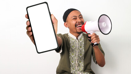 Excited Indonesian Muslim man in koko and peci shows a blank phone screen and holds a megaphone,...