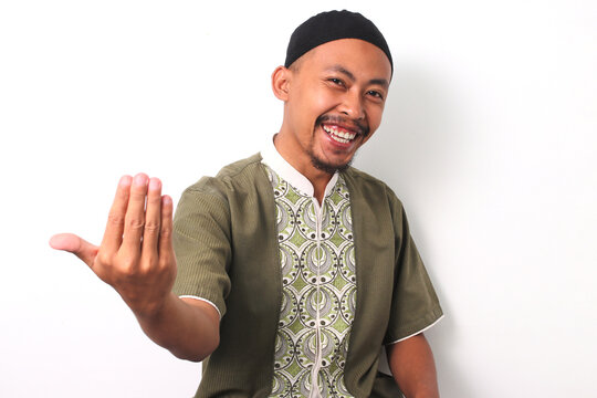 Indonesian Muslim man in koko shirt and peci smiles and extends a friendly hand gesture towards the camera, offers a welcoming gesture while standing on white background