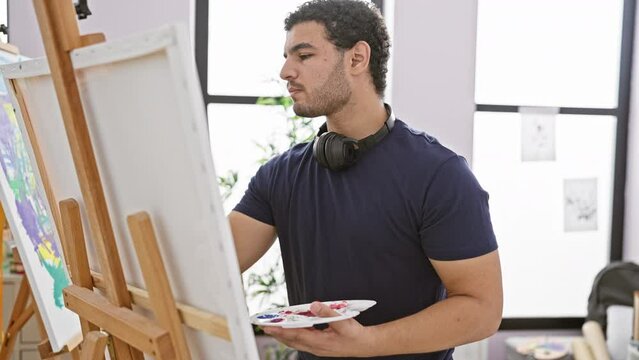 Pensive man with a beard painting on a canvas in a bright art studio, wearing headphones