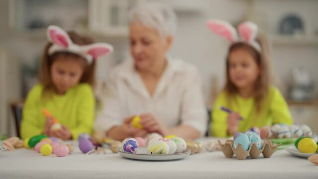 Easter grandmothers with granddaughters. Defocus grandmother with twins grandchildren painting decorating eggs in rabbit bunny ears, talking, celebrating together at home. Easter holiday concept.
