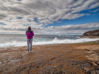 Teenager girl looking at stunning nature scenery with ocean and rough stone shore and cloudy sky....
