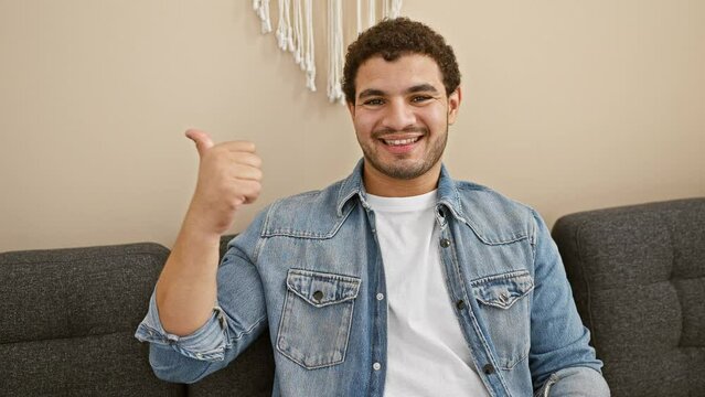 Cheerful young arabian man with beard, joyfully pointing finger sideways, giving thumbs up from sofa in his home interior with open-mouth smile