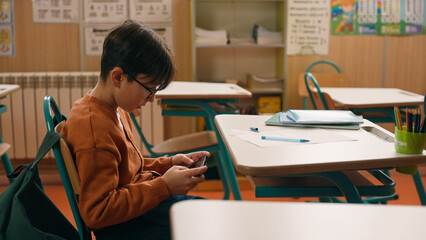 Child kid son pupil student bad behavior boy schoolboy playing video game on mobile phone at lesson at school hide smartphone under class desk distract from studying gadget addiction writing learning - obrazy, fototapety, plakaty