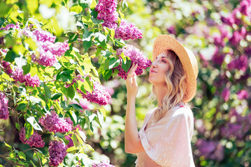 Young Woman In Summer Hat Enjoying The Fragrance Of Lilac Blooms On Sunny Day. lady in a straw hat and dress smelling lilac flowers in a sunlit garden. - 752966074