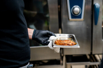 chef in black attire and gloves holds tray of grilled meat next to a stainless steel commercial oven. The grilled meat has visible grill marks, and the person stands in an industrial kitchen setting.